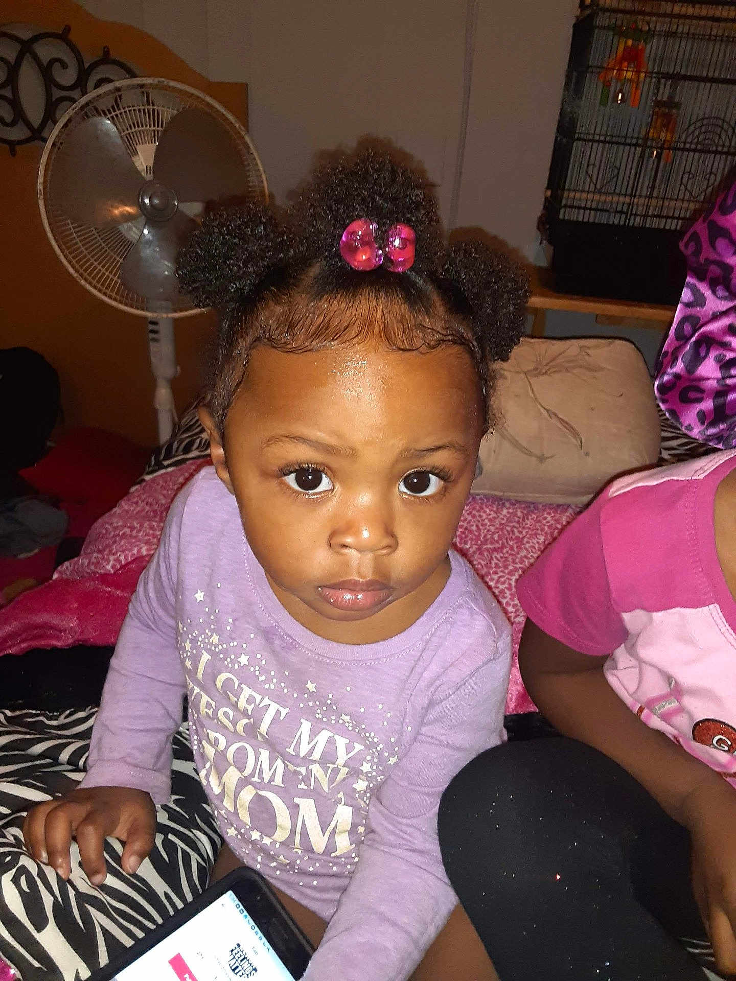 Za'Mierra Grace is registered to the contest to win money with this photo: toddler, child, phone, bed, patterned_bedspread, hair_puffs, purple_shirt, indoor, fan, birdcage, curious, person, face, eyes, hand, sitting, clothing, furniture, pink, black_pants