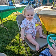 Phoenixx is registered to the contest to win money with this photo: adult_hand, balloons, celebration, child, clothing, curly_hair, fence, folding_chair, grass, green_tablecloth, happy, outdoor, party, person, photograph, plate, snack, sunlight, table, toddler