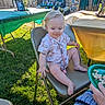 adult_hand, balloons, celebration, child, clothing, curly_hair, fence, folding_chair, grass, green_tablecloth, happy, outdoor, party, person, photograph, plate, snack, sunlight, table, toddler