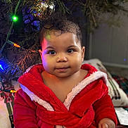 Rose is registered to the contest to win money with this photo: baby, child, red_robe, christmas_tree, christmas_lights, ornament, holiday, indoor, cute, smiling, face, person, festive, decorations, soft_lighting, blanket, cozy, warm, portrait, holiday_season
