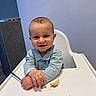 baby, bread, chair, child, crumbs, cute, face, feeding, hands, happy, highchair, indoor, infant, mealtime, portrait, shirt, smile, spoon, table, wall