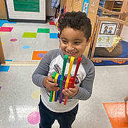 Justin joined the competition — help win amazing prizes! child, smiling, toy, classroom, colorful_floor, educational_posters, happy, standing, curly_hair, casual_clothing, indoor, playful, young_boy, blue_shoes, wooden_furniture, school_environment, early_learning, bright_colors, floor_tiles, holding_toy