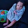baby, child, tongue_out, striped_blanket, bucket, candy, toys, blue_bucket, gray_clothing, sitting, playful, cute, dark_background, indoor, infant, person, smiling, legs, hand, face