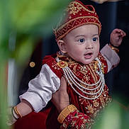 Nayan is registered to the contest to win money with this photo: baby, child, traditional_clothing, red_outfit, gold_embroidery, pearl_necklaces, hat, portrait, hand, celebration, festive, expression, wide_eyes, indoor, decorative, cultural, person, closeup, smile, jewelry