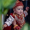 baby, child, traditional_clothing, red_outfit, gold_embroidery, pearl_necklaces, hat, portrait, hand, celebration, festive, expression, wide_eyes, indoor, decorative, cultural, person, closeup, smile, jewelry