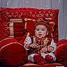 baby, child, red_velvet, throne, traditional_clothing, hat, necklace, gold_embroidery, jewelry, couch, portrait, indoors, cute, drool, hands, smile, sitting, luxury, decoration, fabric