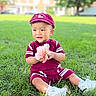 cap, casual_clothing, child, cute, daylight, flower, grass, greenery, happy, maroon_outfit, nature, outdoor, playful, portrait, sitting, smiling, summer, toddler, white_shoes, young_child