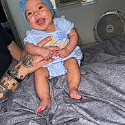 Hyacinth is registered to the contest to win money with this photo: baby, child, smiling, blue_bow, headband, tattoos, bed, blanket, indoor, fan, air_conditioner, person, infant, clothing, happy, holding, skin, foot, hand, room