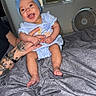 baby, child, smiling, blue_bow, headband, tattoos, bed, blanket, indoor, fan, air_conditioner, person, infant, clothing, happy, holding, skin, foot, hand, room