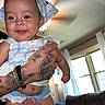 baby, ceiling_fan, child, clothing, curtain, cute, furniture, hand, happy, headscarf, indoor, infant, light, living_room, person, smile, sofa, table, tattoo, window