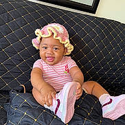 Jnijah joined the competition — help win amazing prizes! baby, child, smiling, pink_hat, striped_onesie, shoe, couch, black_quilted_couch, cute, happy, indoor, playful, sitting, tongue_out, crocheted_hat, footwear, infant, person, portrait, home
