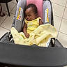 baby, sleeping, stroller, blanket, yellow_clothing, indoor, tile_floor, pink_cushion, black_chair, red_rug, peaceful, infant, child, person, cute, cozy, portrait, resting, curly_hair, car_seat