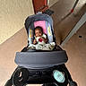 baby, stroller, infant, smile, child, baby_carrier, drink_bottle, person, concrete_floor, wall, door, handlebar, baby_clothing, indoors, floor_mat, seat, blanket, happy, portrait, cute