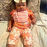baby, bow, child, couch, curly_hair, cute, expression, floral_pants, footwear, indoors, infant, orange_clothing, patterned_pillow, person, pillow, pink_bib, portrait, sitting, skin, white_shoes