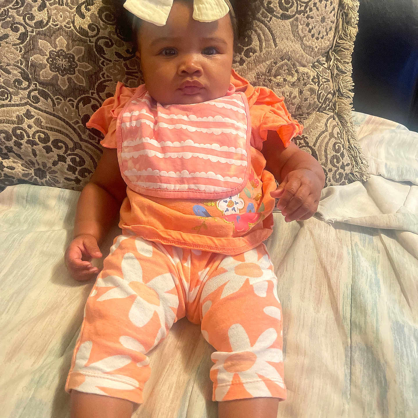 Julianna Benton is registered to the contest to win money with this photo: baby, bow, child, couch, curly_hair, cute, expression, floral_pants, footwear, indoors, infant, orange_clothing, patterned_pillow, person, pillow, pink_bib, portrait, sitting, skin, white_shoes