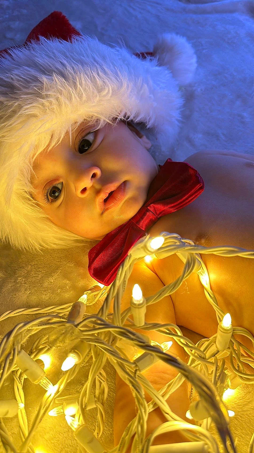Jalei is registered to the contest to win money with this photo: adorable, baby, celebration, child, christmas_lights, close_up, cute, decorations, face, festive, glowing_lights, holiday, indoors, infant, laying_down, portrait, red_bow_tie, santa_hat, soft_blanket, warm_lighting