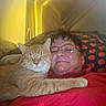 cat, orange_cat, person, woman, face, smiling, glasses, indoors, bed, couch, blanket, red_shirt, cuddle, sleepy, relaxed, whiskers, fur, paw, polka_dot, warm_lighting