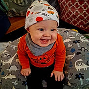 Theodore is registered to the contest to win money with this photo: baby, baby_bib, child, clothing, cushion, cute, decor, dinosaur_pattern, face, halloween, happy, hat, home, indoor, infant, joyful, orange_sweater, playful, seated, smile