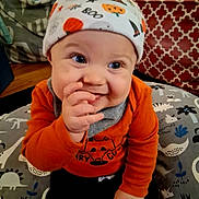 Theodore joined the competition — help win amazing prizes! baby, child, smiling, hat, orange_clothing, hand, face, blue_eyes, indoor, cushion, pattern, dinosaur, happy, cute, infant, toddler, home, cozy, portrait, clothing