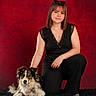 woman, person, dog, pet, portrait, studio, red_background, black_clothing, sitting, kneeling, long_hair, bangs, sunglasses, necklace, tattoo, fluffy, mixed_breed, collar, rug, indoor