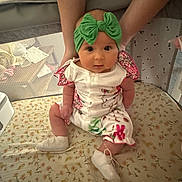 Nora is registered to the contest to win money with this photo: baby, infant, green_bow, headband, floral_outfit, white_shoes, hands, mattress, patterned, nursery, child, cute, sitting, indoor, person, soft_lighting, curious, support, closeup, face