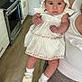 baby, child, dress, bonnet, white_shoes, socks, kitchen, floor, oven, cabinet, person, holding, infant, cute, indoor, flooring, appliance, hand, footwear, clothing