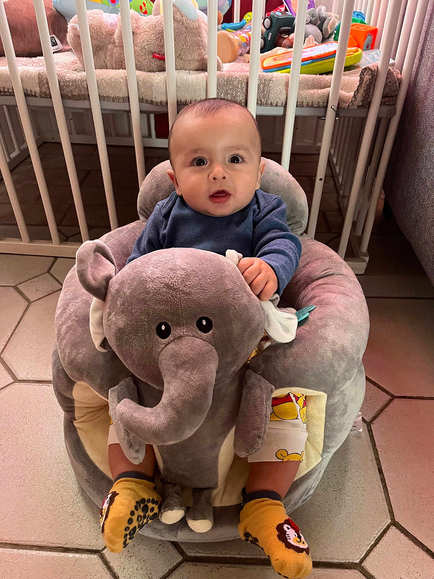 Maé a rejoint le concours — aidez-le/la à gagner de superbes lots ! baby, child, plush_toy, elephant, socks, yellow, nursery, crib, toy, floor_tiles, clothing, cute, indoor, infant, seated, soft_toy, expression, person, furniture, room