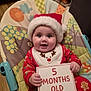 Aleksandra is registered to the contest to win money with this photo: baby, bib, chair, child, christmas, cute, decorations, face, festive, happy, holiday, indoor, infant, pajamas, person, red, santa_hat, sign, smiling, wood_floor