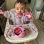 Addison is registered to the contest to win money with this photo: baby, high_chair, messy, food, purple, teether, kitchen, floor, wood, plastic_bowl, child, foot, sitting, face, clothing, expression, curious, indoor, table, tray