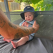 Feonix is registered to the contest to win money with this photo: baby, child, hat, bucket_hat, tattoo, arm, outdoor, cushion, gray_cushion, greenery, plants, fence, wooden_fence, striped_outfit, feet, hands, person, summer, portrait, happy