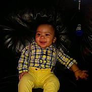 Jeremiah is registered to the contest to win money with this photo: baby, child, infant, smiling, happy, portrait, sitting, plaid_shirt, yellow_pants, suspenders, clothing, leather_chair, indoor, dark_background, blue_bottle, face, eyes, hands, casual, photography