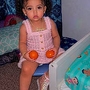 Jaylynn is registered to the contest to win money with this photo: toddler, child, girl, curly_hair, pink_bows, pink_dress, sneakers, oranges, chair, bed, bedroom, carpet, curtains, jewelry, necklace, bracelet, indoor, cute, portrait, playful