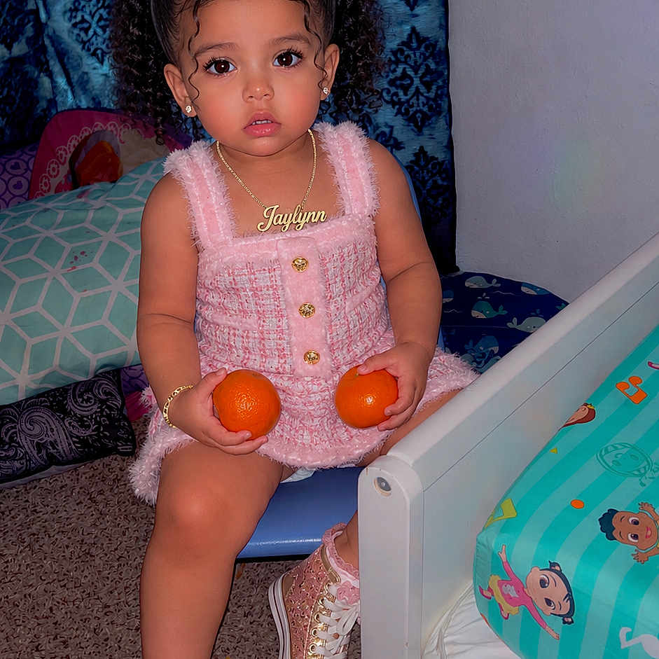 Jaylynn is registered to the contest to win money with this photo: toddler, child, girl, curly_hair, pink_bows, pink_dress, sneakers, oranges, chair, bed, bedroom, carpet, curtains, jewelry, necklace, bracelet, indoor, cute, portrait, playful