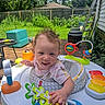 Serena is registered to the contest to win money with this photo: baby, child, activity_center, toy, outdoor, deck, greenery, happy, curly_hair, playtime, sunlight, smiling, person, plastic_toy, colorful, garden, fence, summer, candid, infant