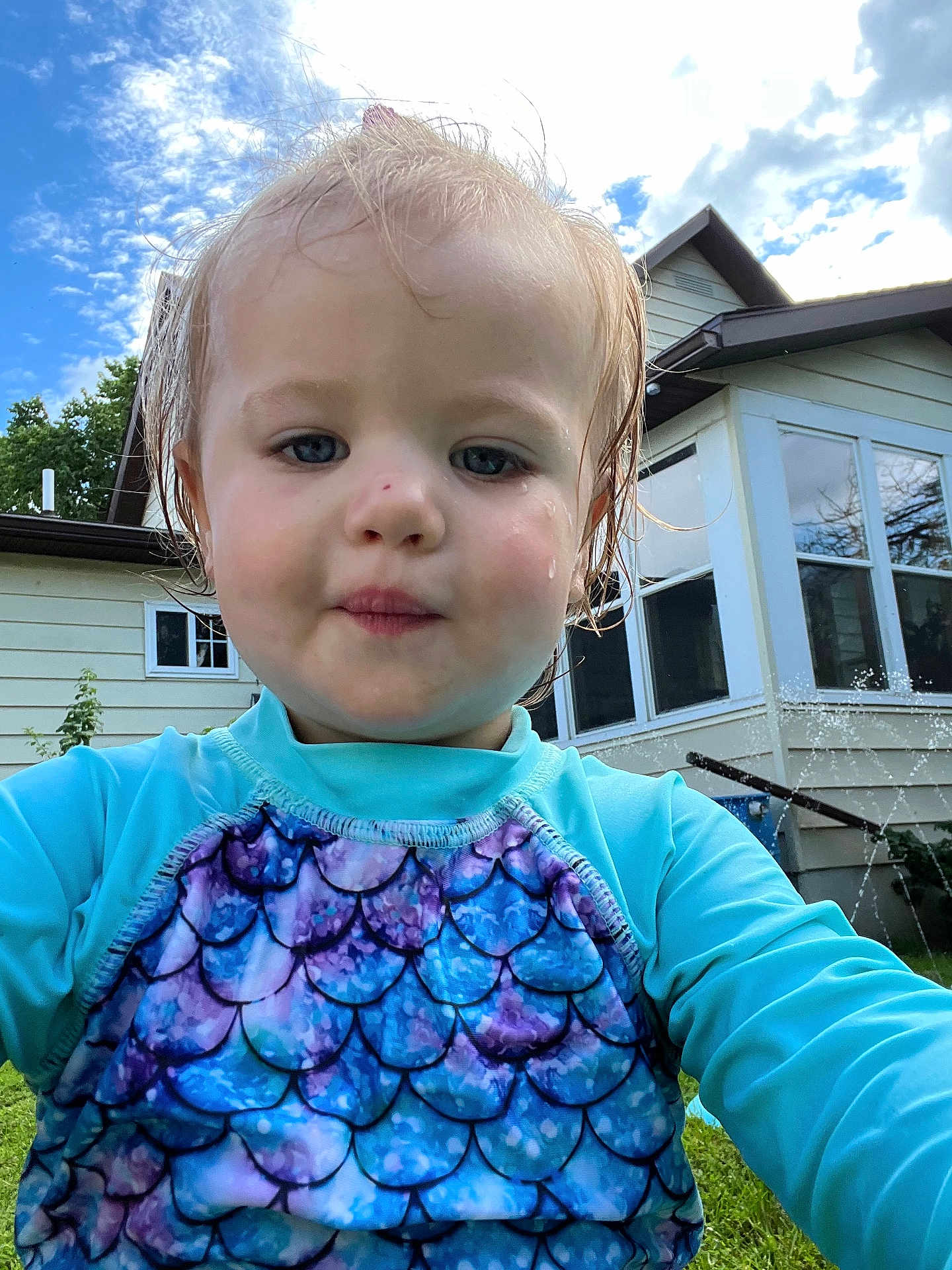 Lyndi is registered to the contest to win money with this photo: toddler, child, face, portrait, outdoor, sky, clouds, house, window, grass, water_spray, swimsuit, wet_hair, blue, purple, pattern, person, summer, daylight, nature