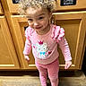 toddler, child, curly_hair, pink_clothing, cat_graphic, polka_dot, barefoot, kitchen_cabinets, wooden_floor, smiling, standing, indoors, cute, happy, person, young_child, cabinet_labels, measuring_spoons, serving_spoons, cutting_boards