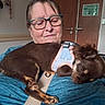 dog, woman, person, sleeping, long_hair, brown_fur, harness, lap_dog, pet, indoors, chair, blue_chair, glasses, closed_eyes, relaxed, cuddling, animal, home, door, accessibility_sign