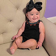 Carmen is registered to the contest to win money with this photo: baby, child, black_bow, smile, sitting, beige_chair, upholstery, purple_wall, cute, infant, happy, clothing, indoor, portrait, headwear, armchair, furniture, skin, person, young