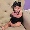 Carmen is registered to the contest to win money with this photo: baby, child, smiling, black_bow, black_outfit, beige_chair, cushion, happy, cute, arm_crossed, indoor, portrait, infant, barefoot, headwear, child_fashion, relaxed, seated, home_interior, purple_wall