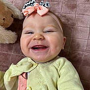 Léhna a rejoint le concours — aidez-le/la à gagner de superbes lots ! baby, smiling, headband, yellow_cardigan, peach_dress, stuffed_animal, soft_blanket, pink_background, happy, cute, child, infant, portrait, indoor, closeup, clothing, baby_girl, headshot, face, playful