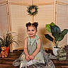 bow, child, curly_hair, cute, decor, dress, floor, girl, happy, home, indoor, plant, portrait, potted_plant, screen, sitting, smiling, teddy_bear, wooden_floor, wreath