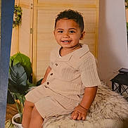 Akique is registered to the contest to win money with this photo: beige_outfit, child, cozy, curly_hair, cute, fluffy_stool, furniture, happy, home, indoor, person, plants, portrait, shirt, shorts, sitting, smiling, toddler, white_sneakers, wooden_background