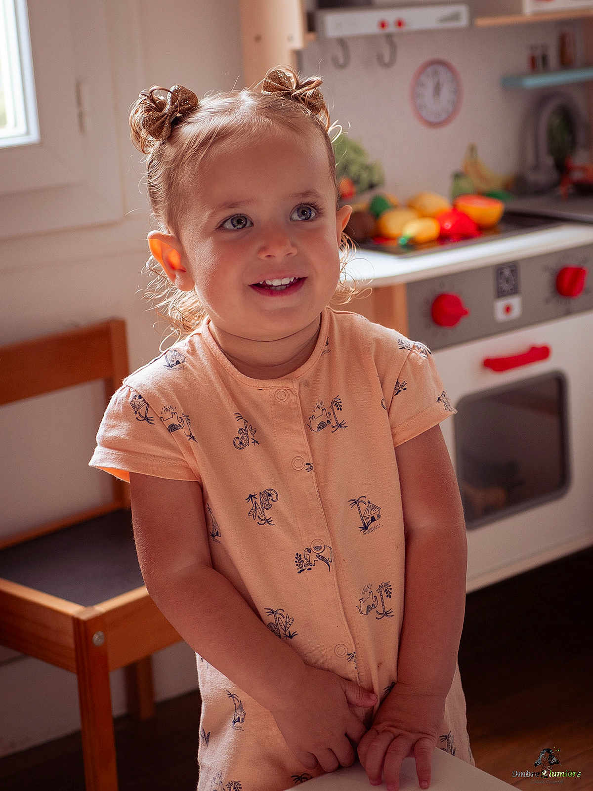 Léonore participe au concours pour gagner de l'argent avec cette photo : child, toddler, smiling, hair_buns, peach_dress, indoor, kitchen, toy_stove, play_food, wooden_chair, window_light, happy, cute, portrait, young_child, playtime, domestic, furniture, person, home