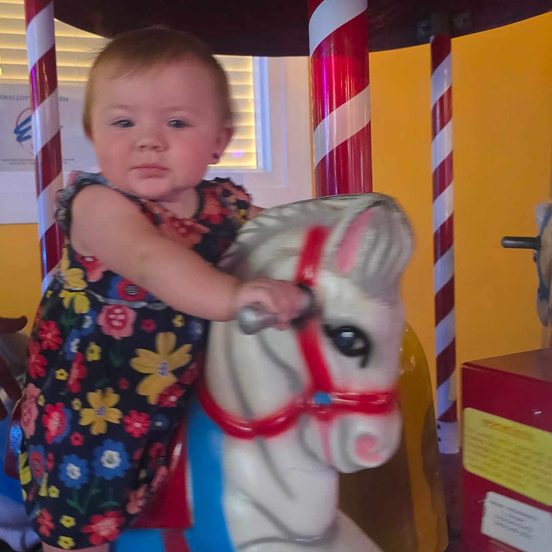 Kelly Kay joined the competition — help win amazing prizes! toddler, child, carousel, horse, ride, flowered_dress, indoor, playful, colorful, candy_striped_pole, yellow_wall, toy, amusement, fun, kid, portrait, cute, baby, face, hand