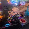 adult_arm, arcade, arcade_floor, car_seat, casual_clothing, child, colorful_lights, entertainment, family, fun, game_machine, gaming, indoor, joy, play, racing_game, smiling, steering_wheel, toddler, young_child