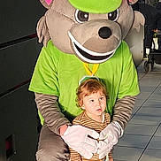 Nao participe au concours pour gagner de l'argent avec cette photo : toddler, child, mascot, costume, indoor, tile_floor, green_shirt, cap, white_gloves, person, footwear, curly_hair, blue_jeans, striped_shirt, holding, posing, cute, playful, large_head, character