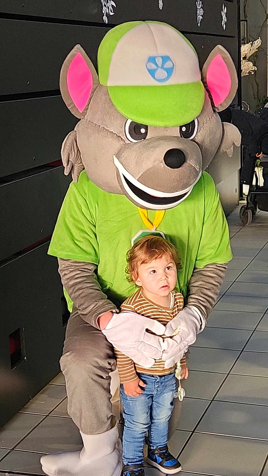 Nao participe au concours pour gagner de l'argent avec cette photo : toddler, child, mascot, costume, indoor, tile_floor, green_shirt, cap, white_gloves, person, footwear, curly_hair, blue_jeans, striped_shirt, holding, posing, cute, playful, large_head, character