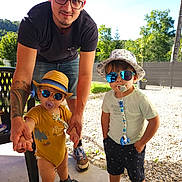 Koda a rejoint le concours — aidez-le/la à gagner de superbes lots ! man, baby, toddler, glasses, sunglasses, hat, pacifier, outdoor, daylight, family, holding_hands, smile, shorts, tshirt, footwear, tattoo, greenery, gravel, chair, person