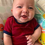 Leo is registered to the contest to win money with this photo: baby, smiling, infant, red_clothing, seated, cushion, patterned_background, animal_print, cute, happy, child, skin, hand, face, eyes, indoor, person, one_person, closeup, young_child