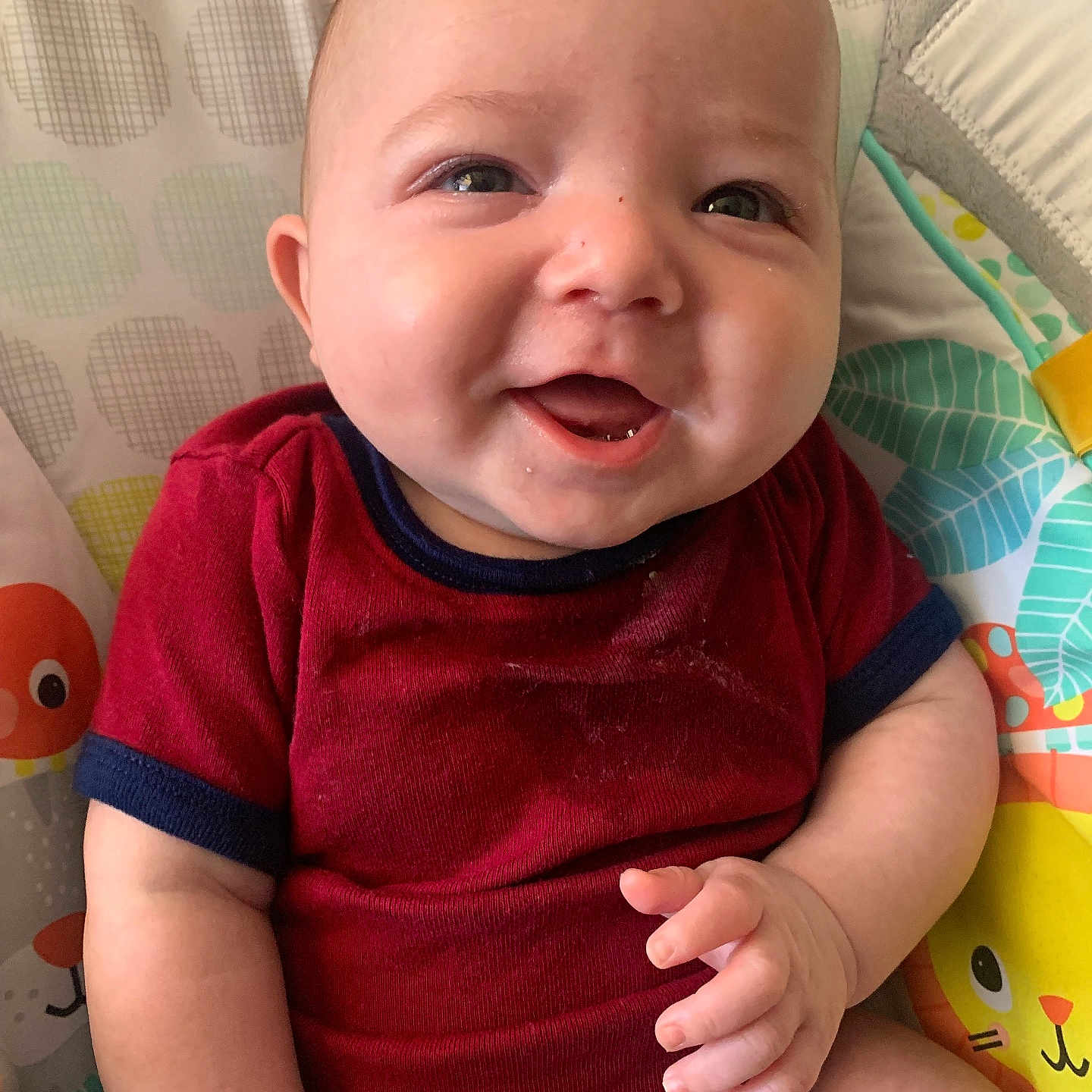 Leo is registered to the contest to win money with this photo: baby, smiling, infant, red_clothing, seated, cushion, patterned_background, animal_print, cute, happy, child, skin, hand, face, eyes, indoor, person, one_person, closeup, young_child